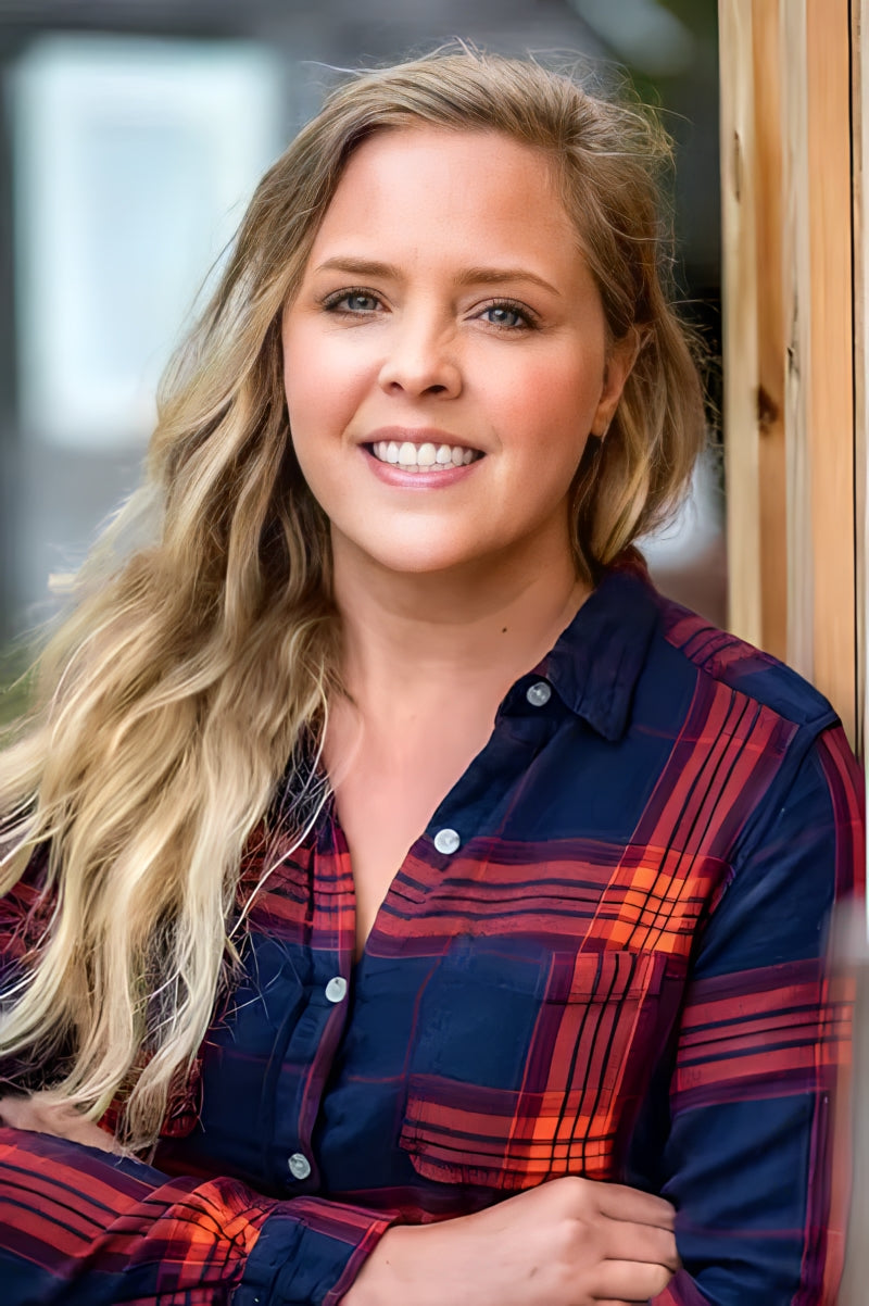 A person with long, wavy blonde hair smiles warmly at the camera. They are wearing a blue and red plaid shirt and leaning against a wooden backdrop. The setting appears to be outdoors, with soft natural lighting.