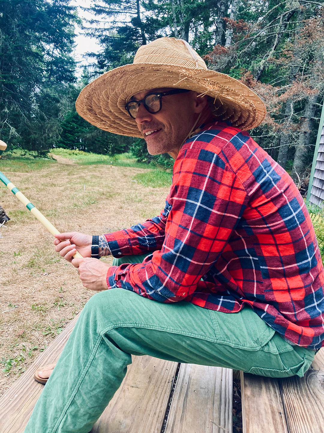 A person wearing a large straw hat, red plaid shirt, and green pants sits on wooden steps holding a croquet mallet. They are outdoors with trees and grass in the background.