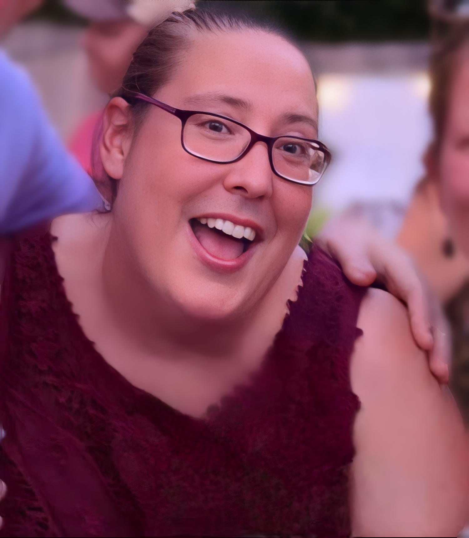 A smiling person with glasses, wearing a burgundy top, is enjoying a moment outdoors. Another person's arm is resting on their shoulder in a gesture of friendship. The background is softly blurred.