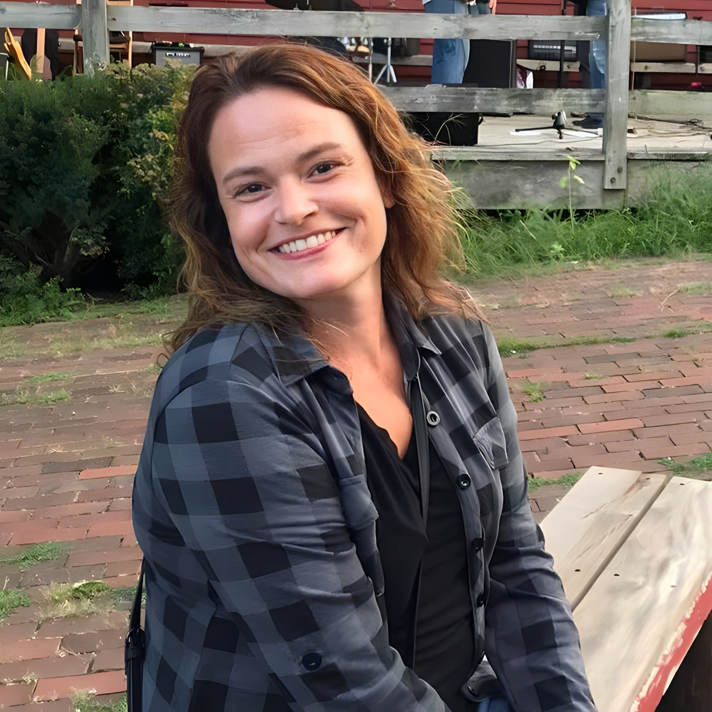 Smiling person with long hair wearing a plaid shirt sits on a bench outdoors. The background includes greenery and a wooden fence.