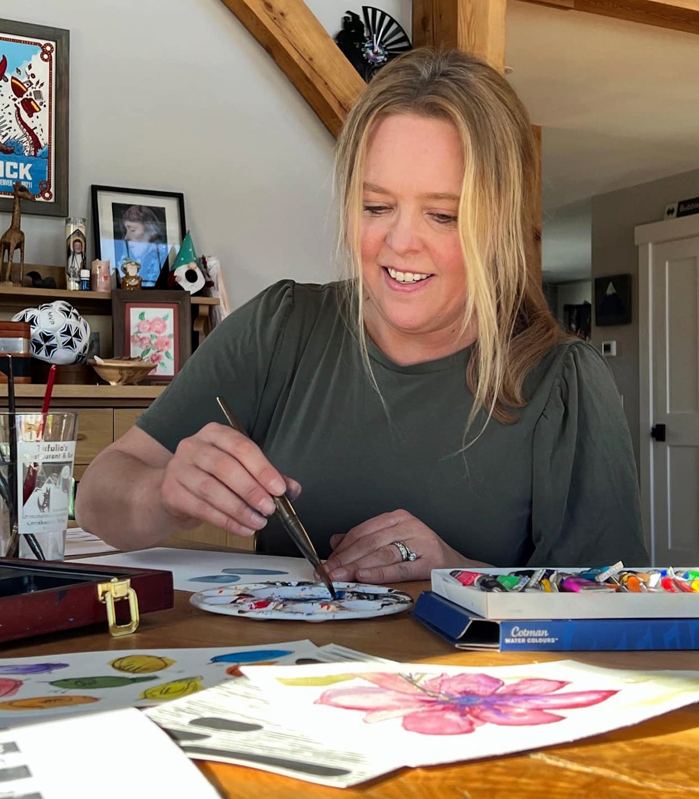 A woman smiles as she uses a paintbrush at a table filled with art supplies and colorful drawings. She is wearing a green shirt in a cozy, well-decorated room with wooden beams and framed art on the walls.