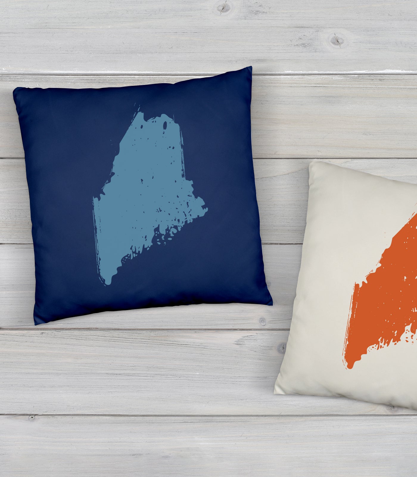 Two decorative pillows on a light wooden surface. The left pillow is navy blue with a light blue abstract shape, while the right pillow is off-white with an orange abstract shape. Both shapes are reminiscent of state outlines.