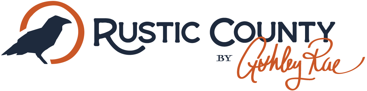 Logo for Rustic County by Ashley Rae, featuring a silhouette of a bird within an orange circular shape to the left of the text. The words "Rustic County" are in dark blue and "by Ashley Rae" is in cursive orange.