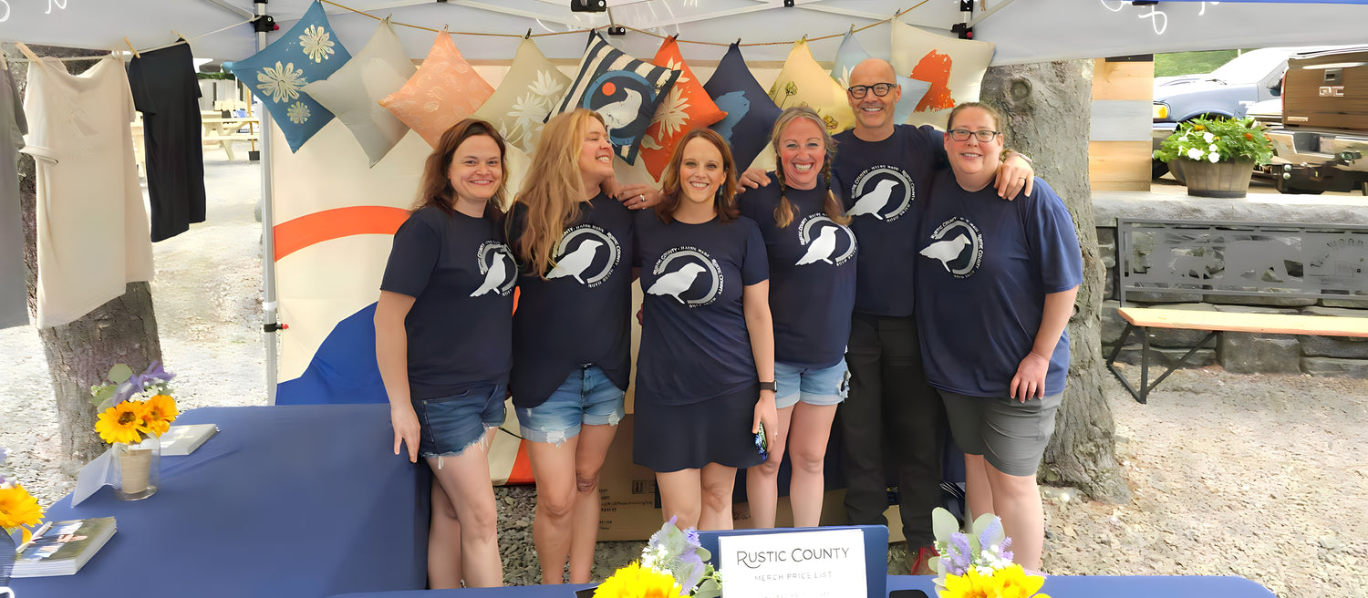A group of six people standing together under a canopy at a booth. They are wearing matching navy blue shirts with a white logo. The booth is decorated with colorful flags and sunflowers, and a sign reads "Rustic County.