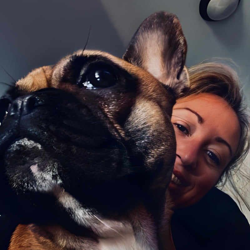 A woman smiling closely behind a French Bulldog. The dog, with its large eyes and alert ears, is in focus while the woman's face is partially visible, showing her joyful expression. The background is softly blurred.