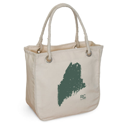 A beige organic cotton Maine Rope Tote from Rustic County with sturdy rope handles and a green brushstroke map of Maine imprinted on one side, along with the initials "RC" in a lower corner.