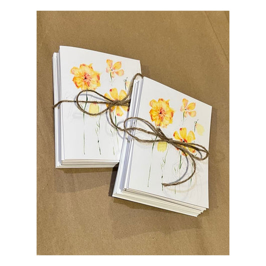 The Watercolor Prints Note Card Bundle by Rustic County is showcased in two stacks, each secured with twine, resting elegantly on a brown surface. These premium paper cards are adorned with watercolor-style orange and yellow flowers on the front, offering a charming touch to any stationery collection.