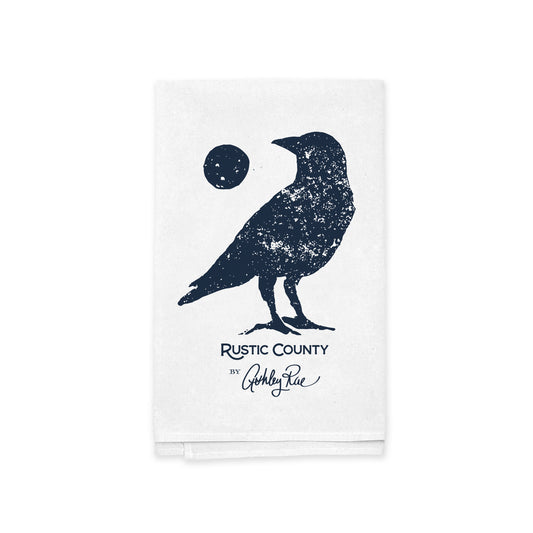 A white Rustic County kitchen tea towel featuring a dark blue Full Moon Crow print of a crow standing beside a small circle, with the text "Rustic County by Ashley Rae" underneath.